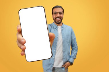 Cheerful indian man in glasses and denim shirt holding a phone with a blank screen, easy to add a custom image clipart