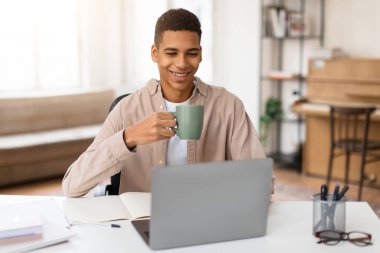 Dizüstü bilgisayarı üzerinde çalışırken bir fincan kahvenin keyfini çıkaran tatmin olmuş genç bir erkek, rahatlık hissi sergiliyor.