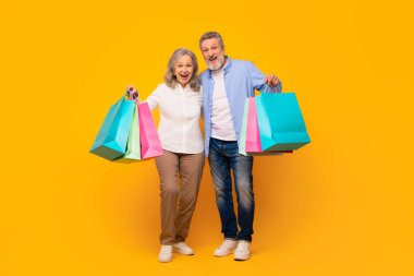Renkli alışveriş çantaları taşıyan mutlu koca ve eş Kara Cuma satışlarını kutluyor, sarı stüdyo arkaplanına karşı alışverişlerle birlikte ayakta duruyor. Olgun tüketim. Tam uzunluk