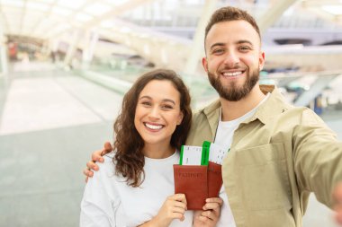 Mutlu çift, havadar ve modern bir havaalanı alanının arka planında pasaport ve biniş kartlarını sergileyerek selfie çeker.