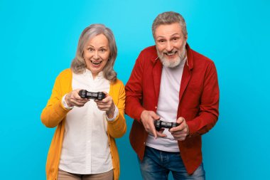 Gündelik bilgisayar oyunlarında mutlu çift dijital eğlence ve eğlencenin tadını çıkarıyor, oyun alanı kontrolörleriyle birlikte mavi stüdyo arka planında duruyorlar, birlikte online oyunu kazanıyorlar.