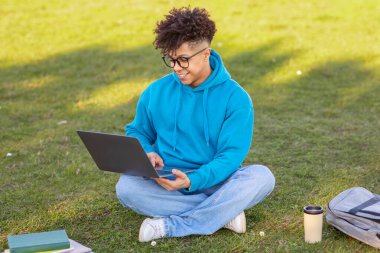 Casual young african american man in a bright blue hoodie sitting cross-legged in a grassy field, working on a laptop with a coffee cup nearby clipart