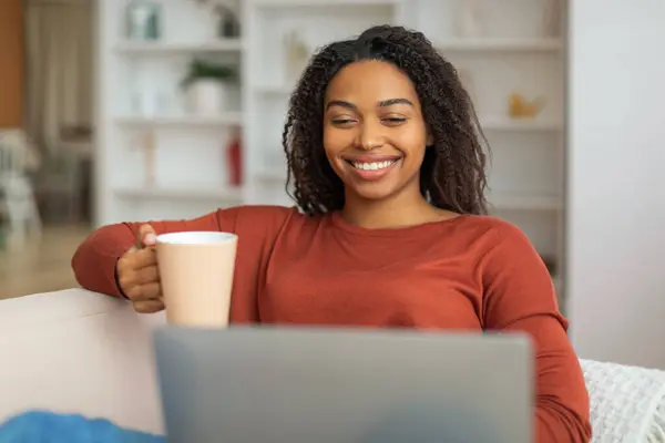 Smiling black woman sitting comfortably on a couch, holding a coffee cup, african american female focused on a laptop screen in front of her.