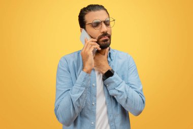 Gözlüklü ve mavi tişörtlü, sakallı bir Hintli doktorla akıllı telefonuyla konuşuyor, boğazına dokunuyor.