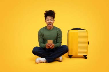A joyful young african american man sitting cross-legged next to a yellow suitcase on a yellow background, holding a smartphone and looking excited clipart