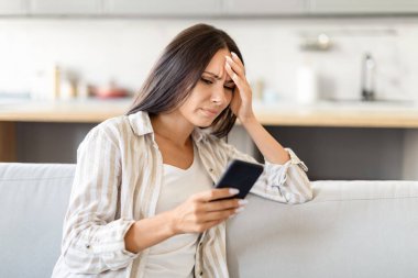 Sıradan giyinmiş bir kadın, koltukta oturan akıllı telefon ekranına bakarken rahatsız ya da endişeli görünür.