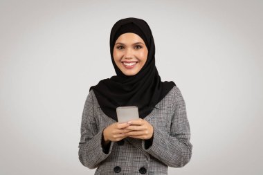 Kendine güvenen Müslüman iş kadını şık gri ekose takım elbiseli ve siyah tesettürlü, mutlu bir şekilde akıllı telefon tutan, modern profesyonel kadınları temsil eden