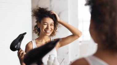 Gülümseyen afro kız saçını banyoda aynanın önünde kurutuyor..