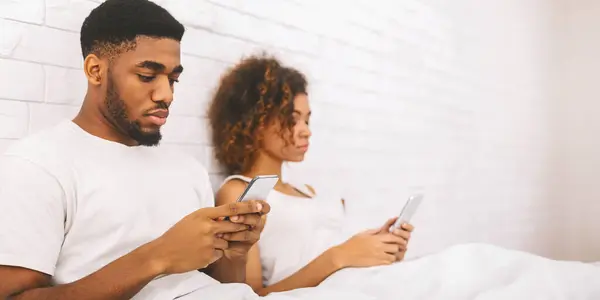 African American couple in bed with back turned and absorbed in their smartphones, reflecting modern relationships
