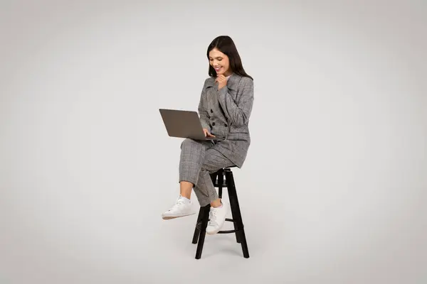 stock image Relaxed and focused young businesswoman browsing on laptop while sitting on chair, displaying casual work style, against light background