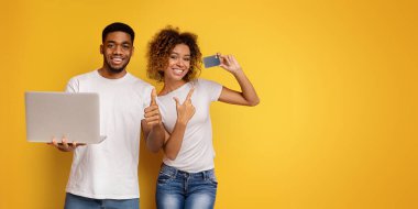 Genç, gülümseyen bir Afro-Amerikan çift sarı bir arkaplanda durur, erkek dizüstü bilgisayarını tutarken kadın başparmağını kaldırır..
