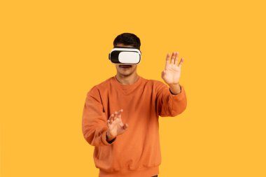 Bir adam sanal gerçekliğe dalmış, VR kulaklık takıyor ve elleri ile canlı turuncu bir arka plana işaret ediyor, yüzü gizlilik için gizlenmiş.