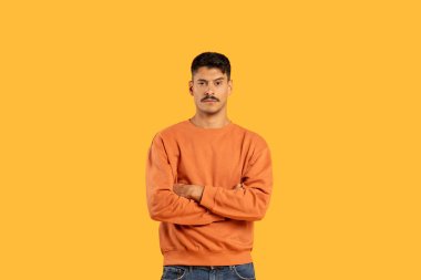 A stern-looking man with moustache stands with arms crossed on a solid yellow background, copy space clipart