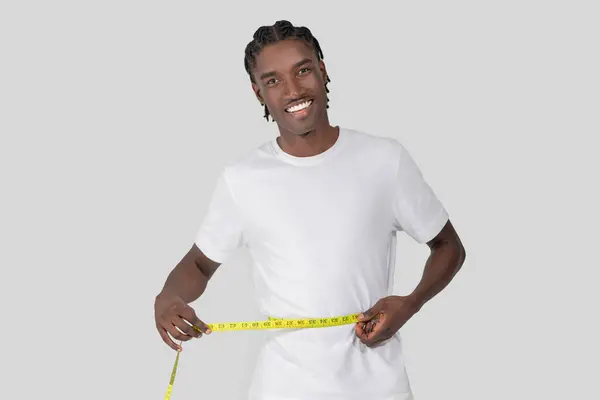 笑顔のアフリカ系アメリカ人男性は誇らしげに腰を測定し 白い背景にフィットネスの進歩と健康を示しています — ストック写真