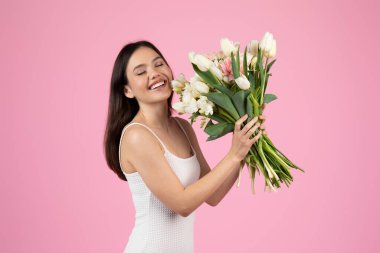 Görüntü, gülen Avrupalı bir bayanı çiçeklerle yazın tadını çıkarırken yakalıyor. Z jenerasyonunun enerjisini somutlaştırıyor ve pembe bir arka planda tek başına duruyor.