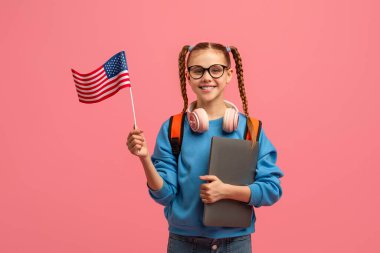 Gözlüklü ve sırt çantalı neşeli kız elinde pembe arka planda bir Amerikan bayrağı tutuyor, internetten İngilizce öğreniyor.