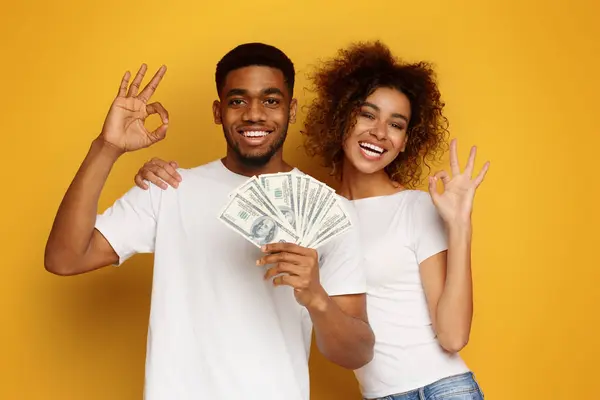 Great investment. Happy african man and woman gesturing OK sign with lots of money, orange studio background