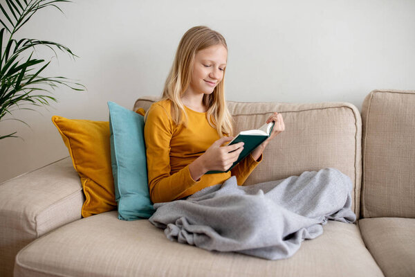 Serene teen girl is engrossed in reading a book while comfortably seated on a couch with cushions and a blanket, copy space