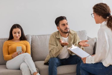 Bir erkek, ortağı koltukta telefonunu kullanırken elinde dosya olan bir kadına kafa karışıklığıyla el kol hareketi yapar.