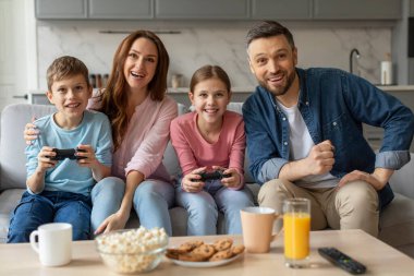Dört kişilik bir aile, oyun kontrolörleri elinde ve gülümseyerek ev içinde video oyunu oynamaktan zevk alır.
