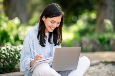 Kendini işine adamış genç bir kız öğrenci bilgisayarını parkta ders çalışmak için kullanıyor, not defterine not alıyor.