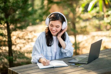 Neşeli kadın bir deftere yazı yazarken müzik dinlemeyi seviyor. Açık hava parkındaki masada dizüstü bilgisayar. Öğrencilerin online dersi var.