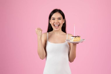 Genç bir kadın bir elinde pasta, diğerinde zafer jesti pembe arka planda heyecan gösterir.