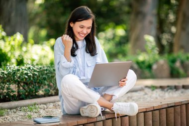 Keyifli genç bir kadın, bilgisayarındaki iyi haberlerden sonra bir başarı jesti sergiliyor. Halk parkında okuyor.