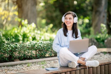Güler yüzlü Avrupalı genç bir kadın güneş ışığında bir bankta otururken kulaklıklarla mutlu bir şekilde yanında akıllı telefonuyla dışarıda bilgisayarla çalışıyor.