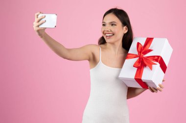 Beyaz elbiseli neşeli bir kadın pembe arka planda kırmızı kurdeleli büyük beyaz bir hediye kutusuyla selfie çeker.