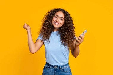 Akıllı telefonu tutan neşeli genç bayan ve sarı parlak arka fonun önünde kazanan bir yumruk pompası yapıyor.