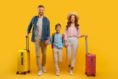 Üç kişilik gülümseyen bir aile, anne-baba ve oğul, sarı arka planda bavullarını tutuyorlar, tatil için hazırlar.
