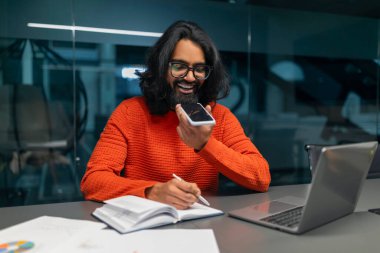 Rahat, genç bir erkek, canlı bir süveter içinde rahat bir ofis odasında telefona doğru konuşuyor.