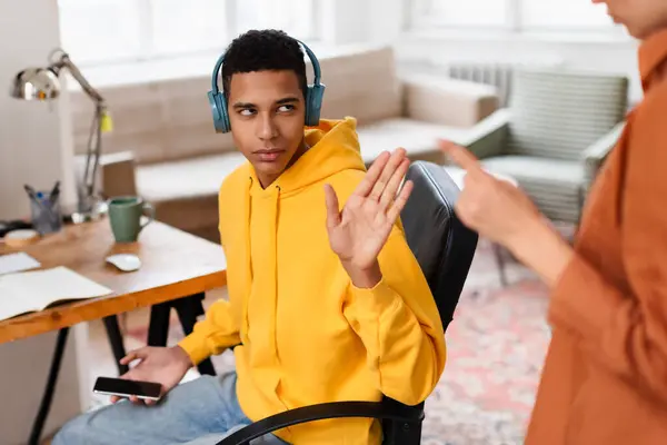 黄色いパーカーとヘッドフォンを着用した若者は 机に座っている間 見えない人にジェスチャーをする — ストック写真