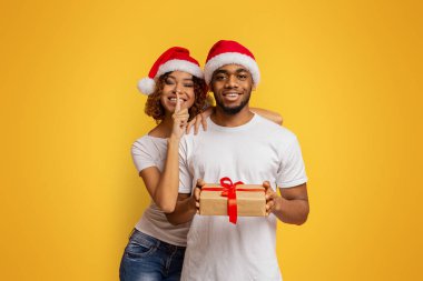 Noel sürprizi. Noel Baba şapkalı Afro-Amerikan çift ellerinde Noel hediyesi, kız sessizlik işareti, turuncu arka plan