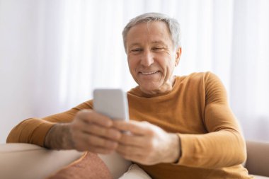 Son sınıf öğrencisi rahat bir erkek oturuyor ve akıllı telefonuyla meşgul. Ev ortamında hoş bir yüz ifadesi sergiliyor.