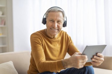 Son sınıf öğrencisi dijital tabletin tadını çıkarıyor ve kulaklıkla ev içi, gülümsemeli bir kulaklıkla ses dinliyor.