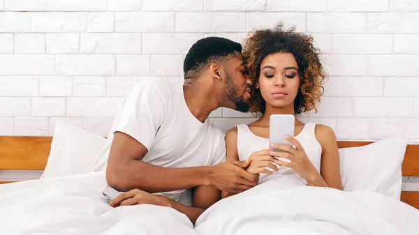 アフリカ系アメリカ人男性は 彼女のスマートフォンに焦点を当てている彼のパートナーにキスしようとします 関係の気晴らしを示しています — ストック写真