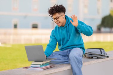 Young brazilian guy student in a blue hoodie sitting on a bench outside with a laptop, books, and backpack, studying or working on a project clipart
