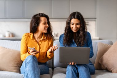 İki gülümseyen kadın kanepede rahatça oturup dizüstü bilgisayarla meşgul oluyorlar, muhtemelen iş tartışıyorlar ya da internette geziniyorlar.