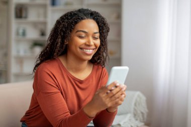 Gülümseyen siyah kadın bir koltukta oturuyor, elinde cep telefonu ekranına odaklanmış, Afro-Amerikan kadın akıllı telefonuyla evde mesajlaşıyor.