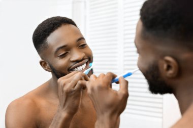 A shirtless African american man brushes his teeth in the mirror, representing good hygiene and personal care habits clipart