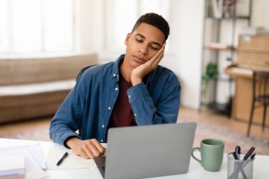 Yorgun bir genç adam, dizüstü bilgisayarla bir masada uykusuzluk belirtileri gösterirken ya kestiriyor ya da gözlerini dinlendiriyor.