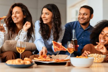 Çok ırklı arkadaşlar, pizza ve şarap dilimleriyle bir yaşam alanı etrafında toplanıyor. Sıcak bir atmosferde yemek ve sohbet paylaşıyorlar.