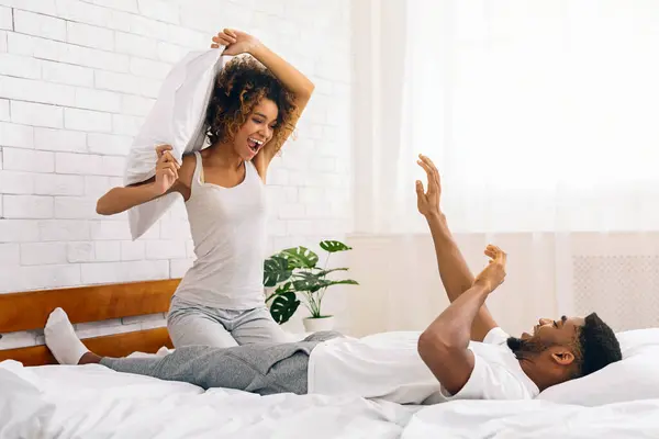 Lighthearted and fun pillow fight scene with a smiling young african american couple on a bed at home