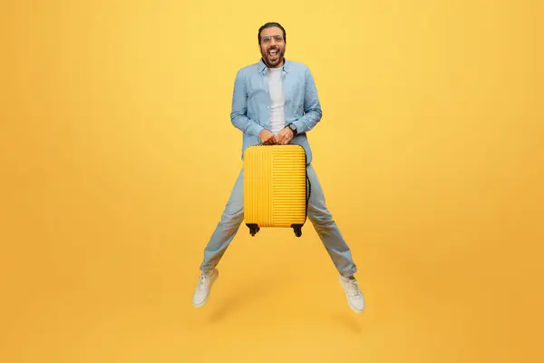 デニム服の幸せなインド人男性は黄色い背景に対して黄色いローリングスーツケースを興奮させます — ストック写真