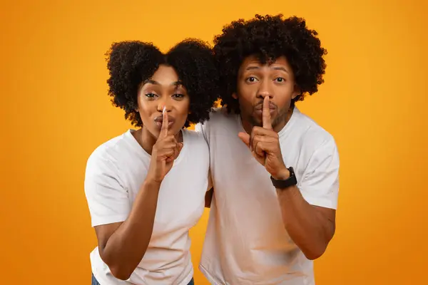 Dont tell anyone. African american man and woman wearing white t-shirts showing silence gesture holding finger on lips, millennial black couple keep quiet, yellow background