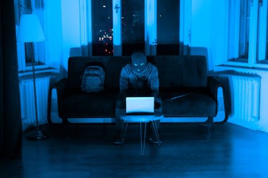 Kar maskeli bir hırsız kanepede oturuyor. Aydınlanmış bir odada bilgi çalmak için dizüstü bilgisayar kullanıyor.