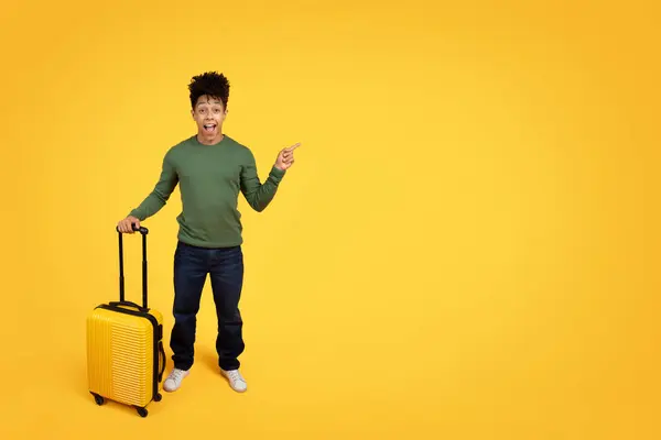 stock image Young excited traveler african american guy pointing to side and holding a carry-on luggage on yellow backdrop