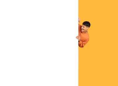 Turuncu kazaklı genç bir adam turuncu arka planda beyaz, dikey, boş bir pankartın arkasından gizlice bakıyor.